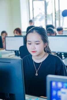 Asian female student focused on a computer in a classroom setting, engaged in study, with classmates in background.